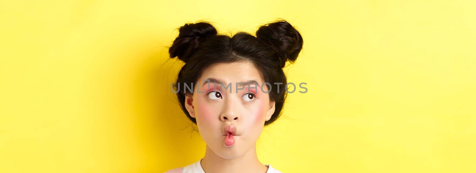 Close-up of teen asian girl pucker lips and looking funny at camera, standing with glamour makeup and stylish hairstyle, yellow background.