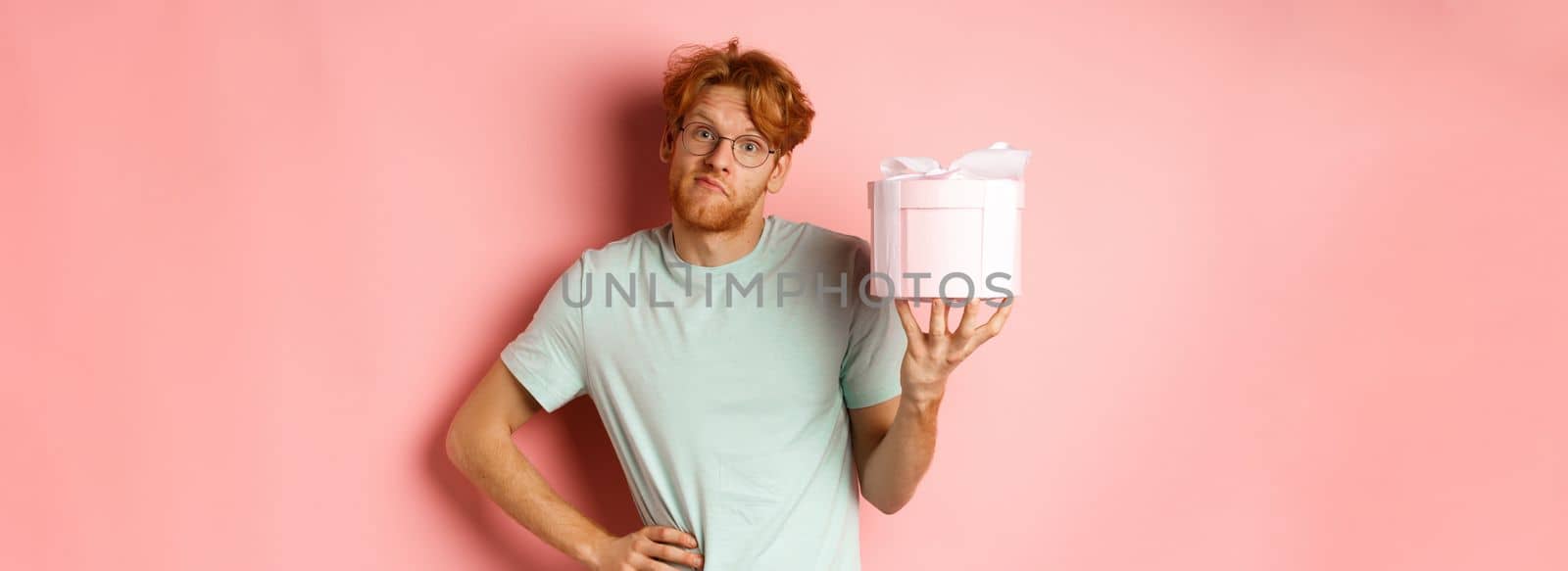Love and holidays concept. Funny redhead guy shrugging silly and showing pink gift box for valentines day, standing over pink background by Benzoix