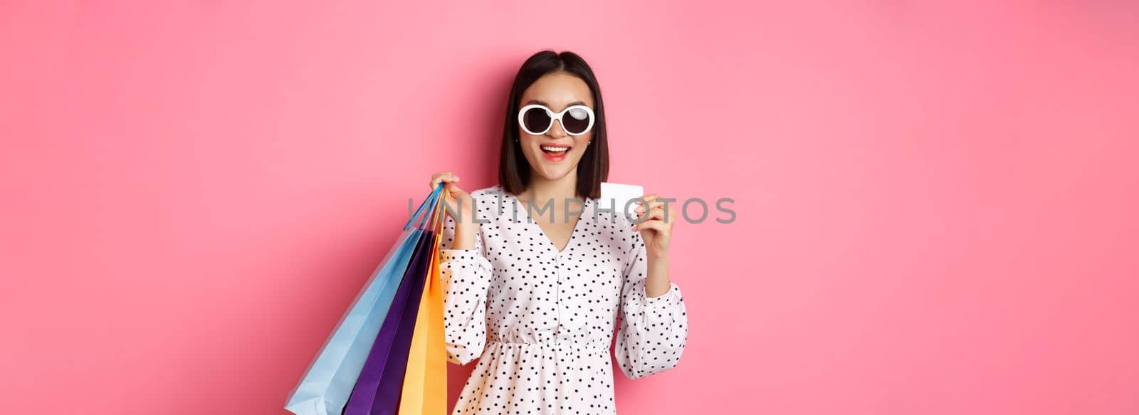 Beautiful asian woman in sunglasses going shopping, holding bags and showing credit card, standing over pink background.