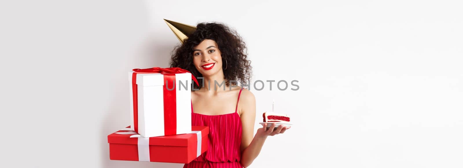 Holidays. Happy elegant woman in party hat and red dress, holding birthday cake and gifts, celebrating b-day, standing over white background by Benzoix