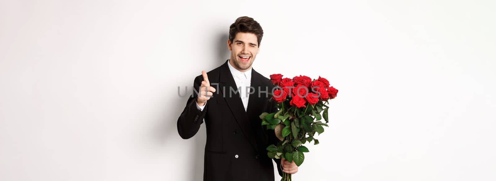 Image of handsome romantic guy in black suit, holding bouquet of roses and pointing at camera, congratulating with holiday, standing against white background by Benzoix