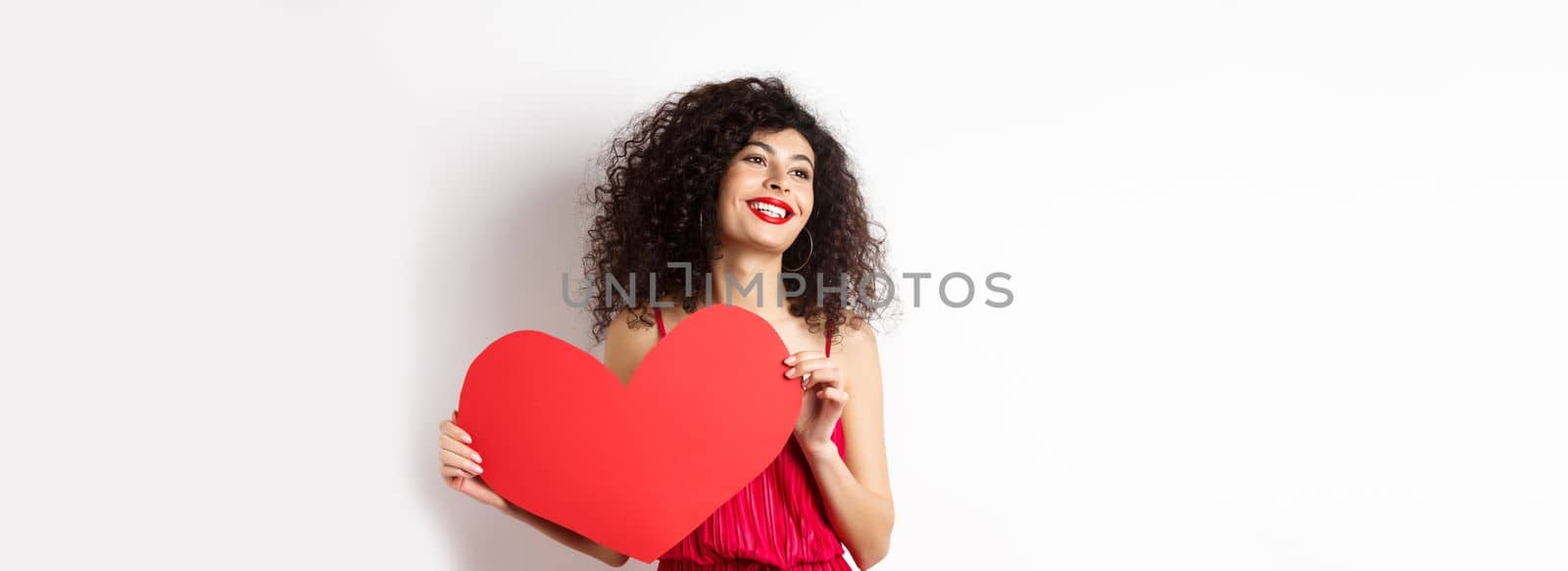Romantic woman in dress and makeup, looking aside with dreamy face and love, showing big red valentines heart card, standing on white background by Benzoix
