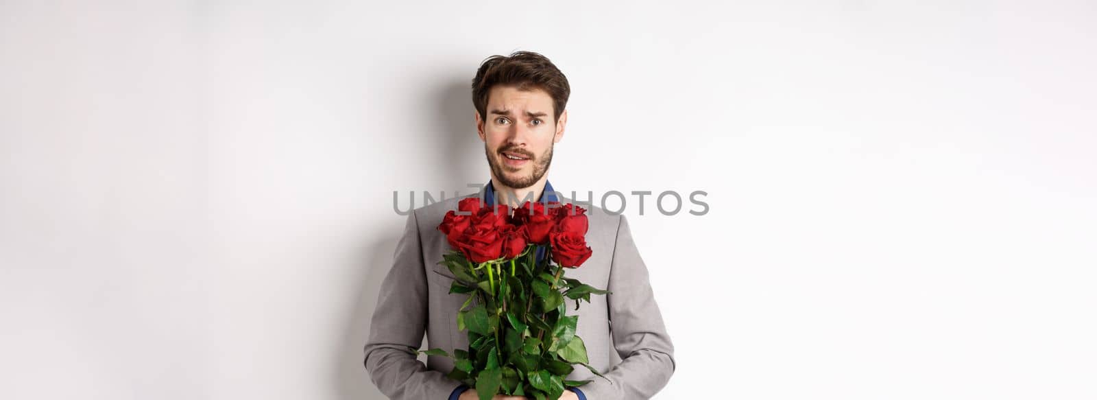 Worried boyfriend in suit, holding flowers roses and looking doubtful at camera, standing with bouquet on valentines day against white background by Benzoix