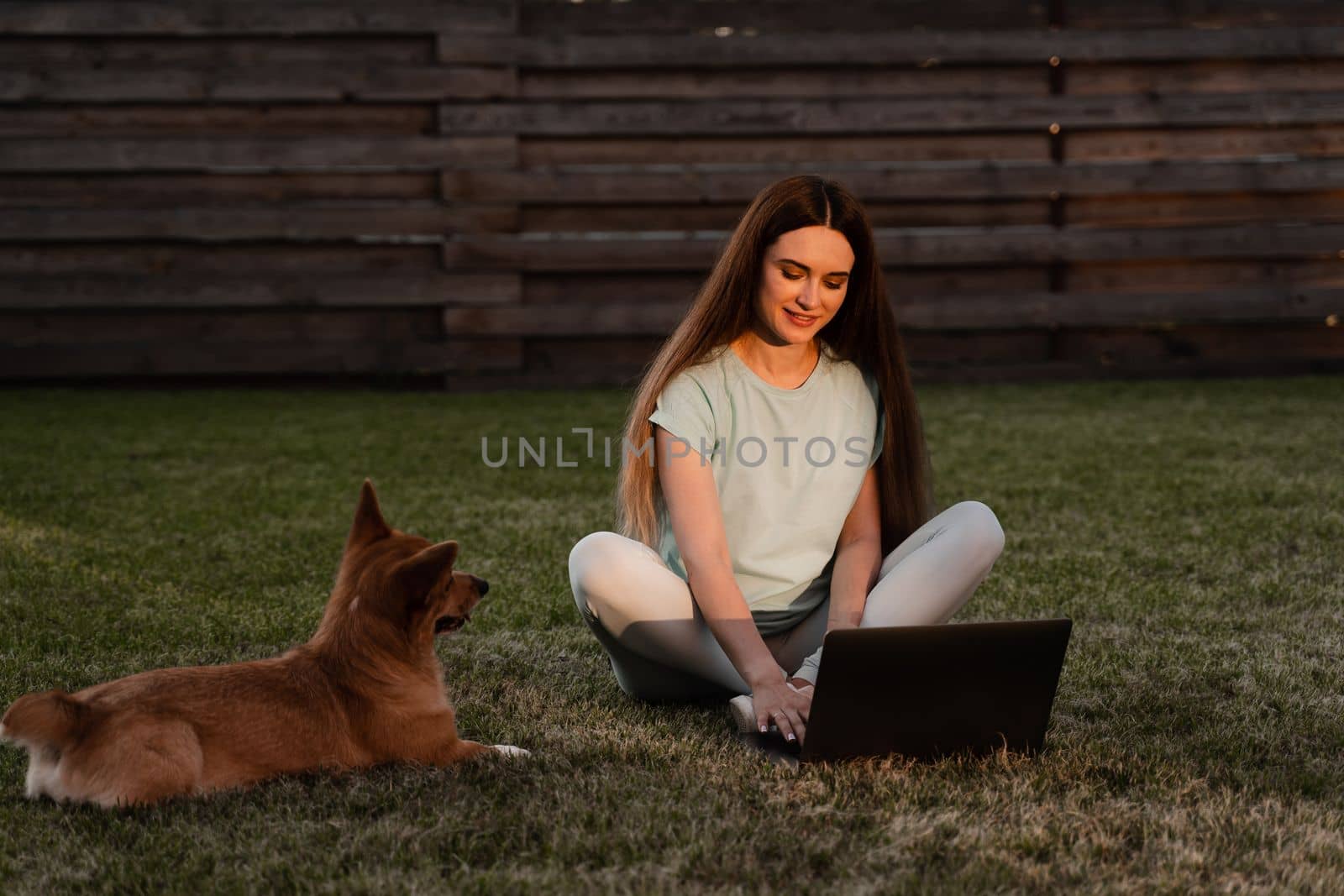 Lifestyle girl and Corgi dog outside. Girl with laptop petting her Welsh Corgi Pembroke dog on the grass. Busy young woman have a break and relax with her dog. Lifestyle with domestic pet. by Rabizo