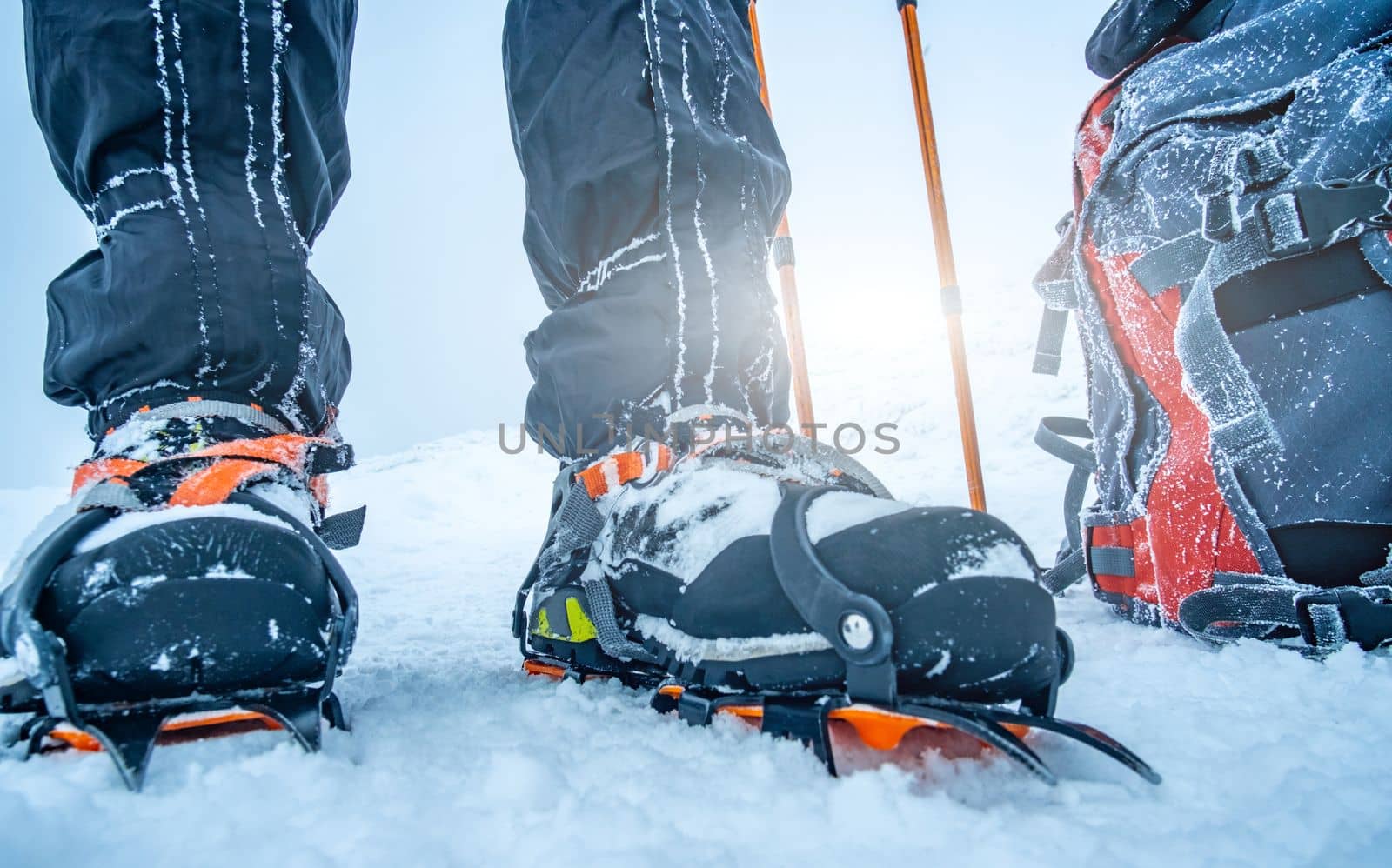 Man wearing crampons standing beside backpack and trekking sticks on deep snow