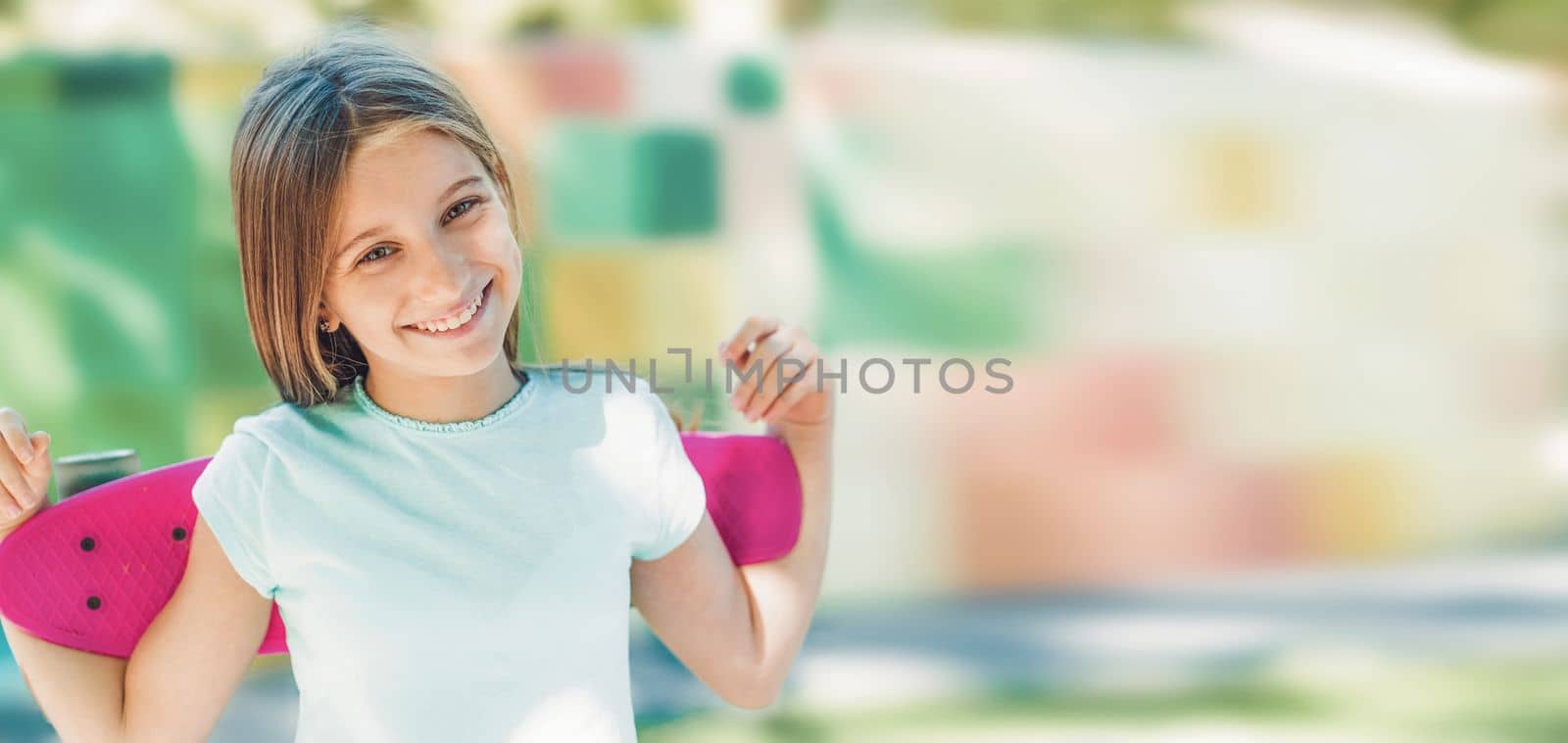 Beautiful girl smiling and holding pink skateboard behind the back. Portrait of teenager with colorful blurred background