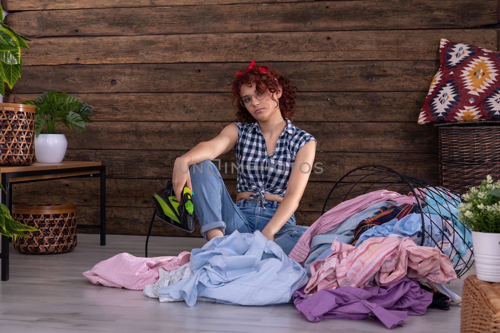 A young girl is waiting to iron men's shirts. She sits on the floor of a room in the house. An overturned basket of clothes. A wall of brown boards.