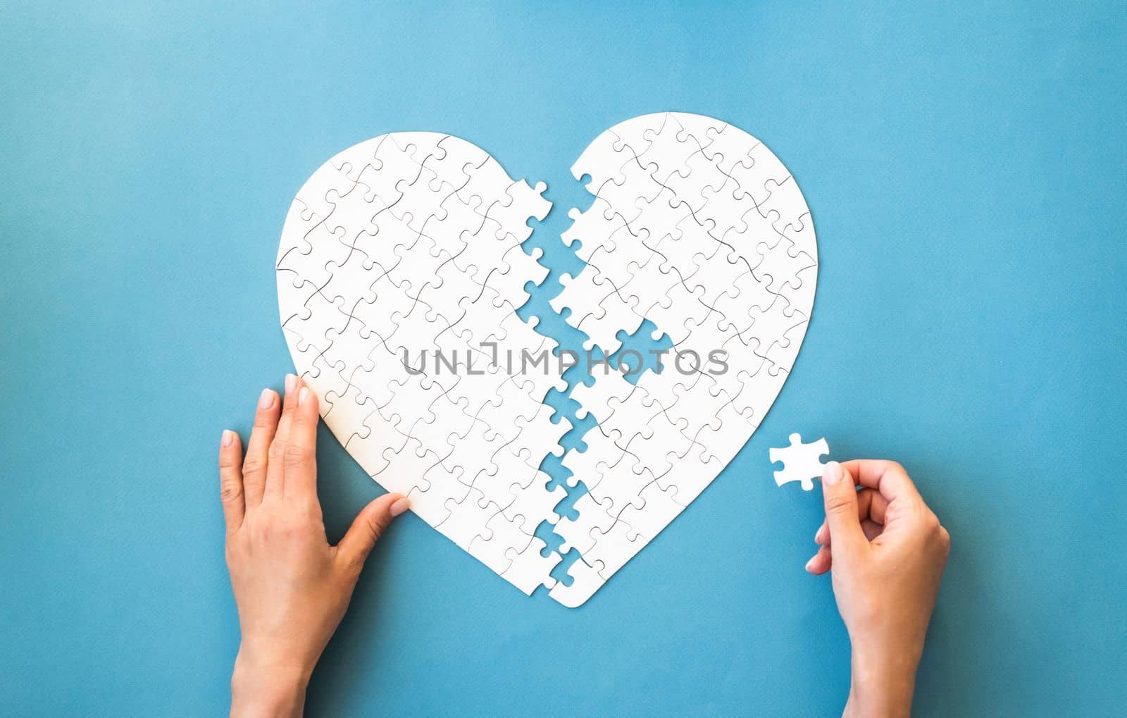 White puzzle in heart shape. Hands connect white details of puzzle on blue background.