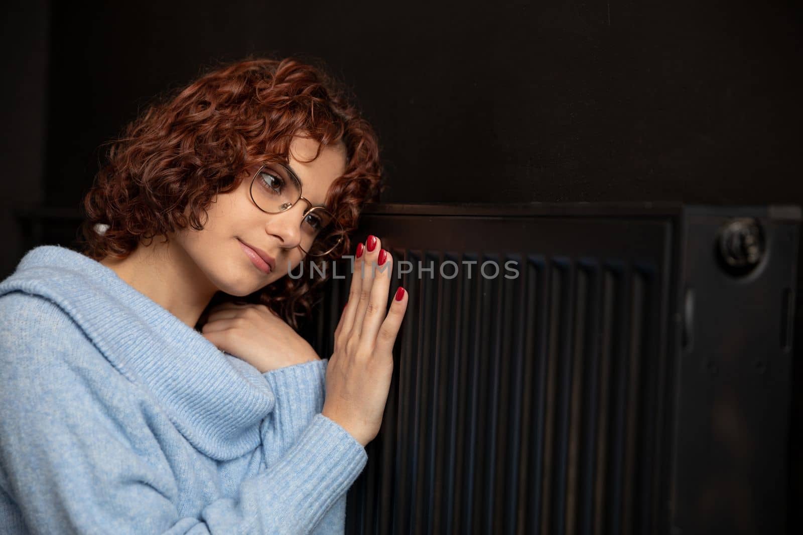 A young girl in a blue thick sweater hugs a warm radiator. Central heating necessary in the house during the winter period.