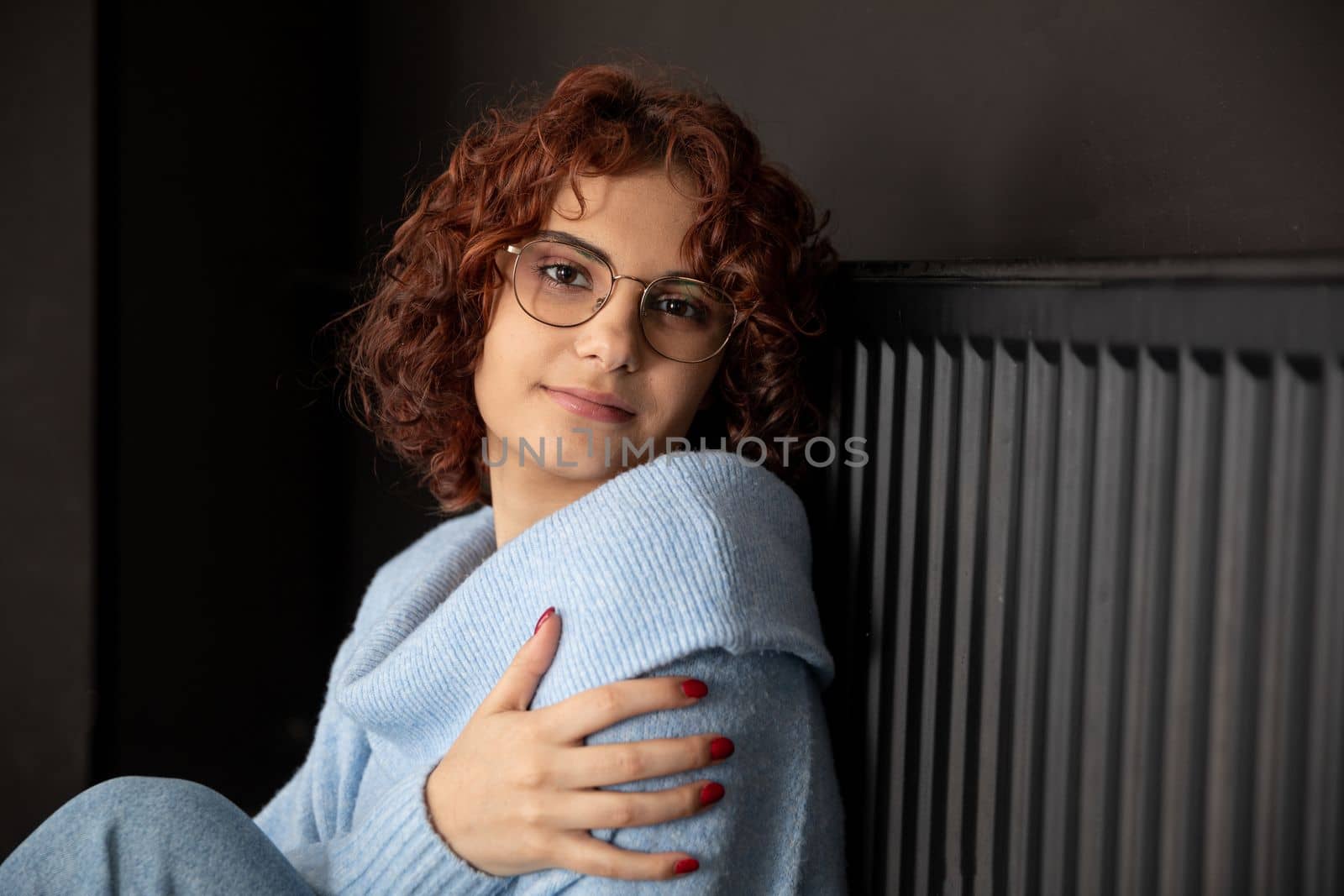 A girl in a blue thick sweater sits by the radiator. Poorly working central heating. A gentle smile.
