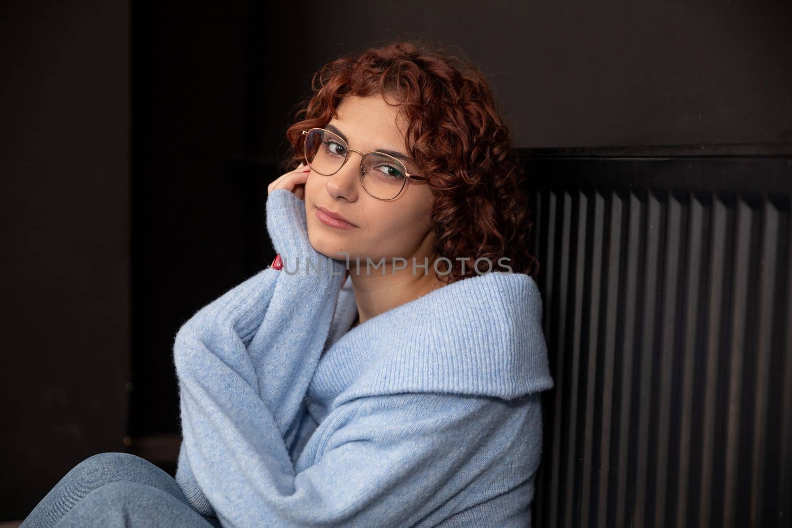 A girl dressed in a thick sweater sits happily by a hot radiator. Heating the rooms of the house with central heating.