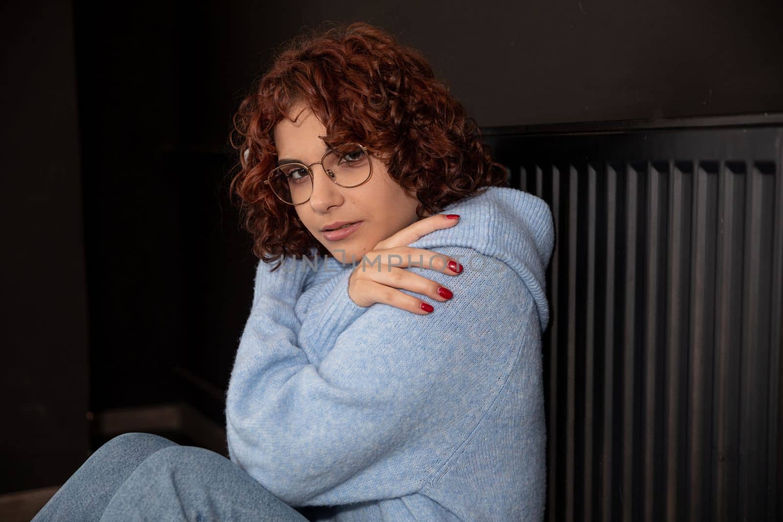A girl in a blue thick sweater sits by the radiator. Poorly working central heating. A gentle smile.