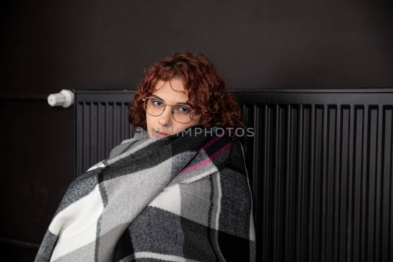 A girl with an imposed scarf leans with her back against a radiator. An attractive woman is freezing in her own home due to a central heating malfunction.