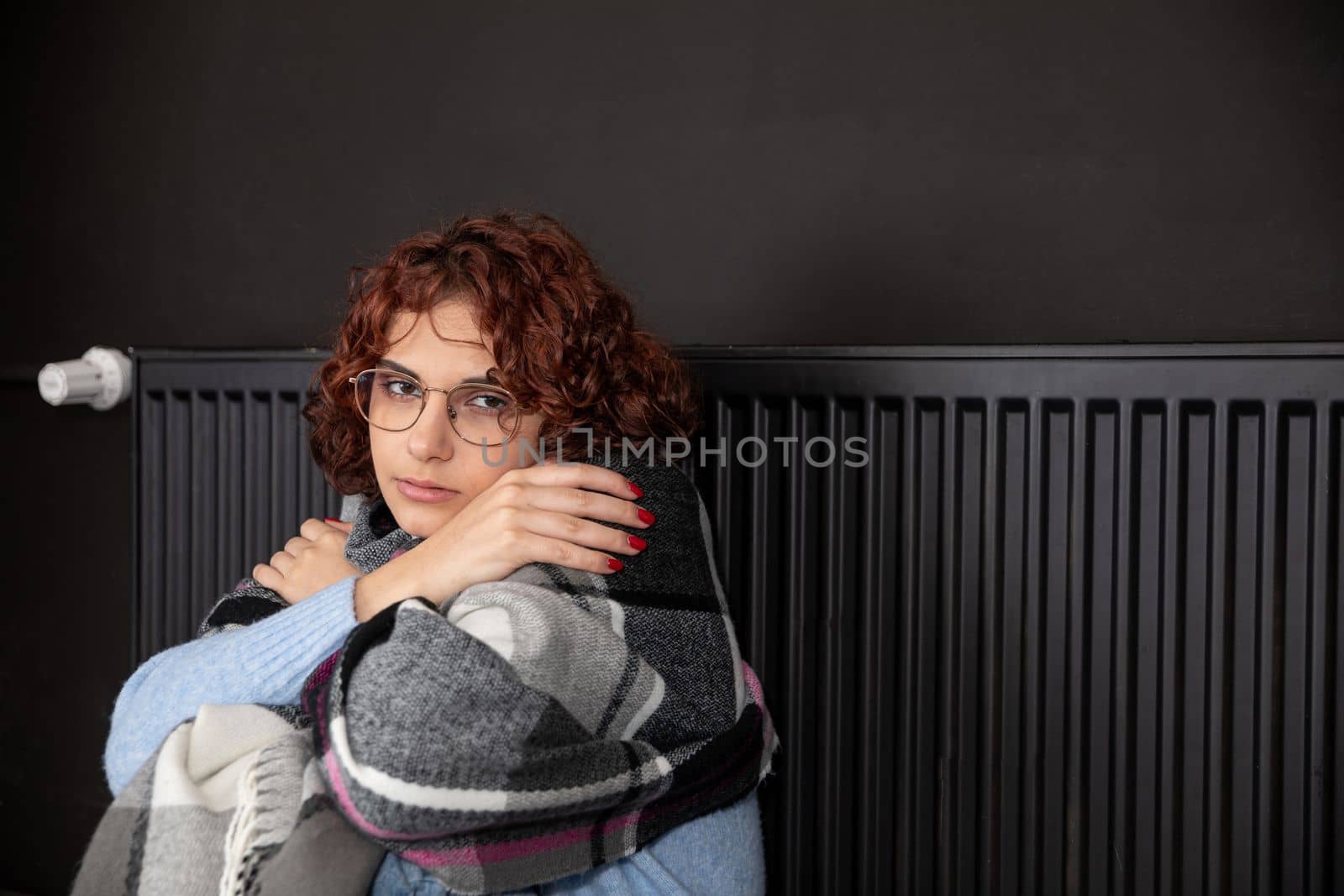 A girl with an imposed scarf leans with her back against a radiator. An attractive woman is freezing in her own home due to a central heating malfunction.