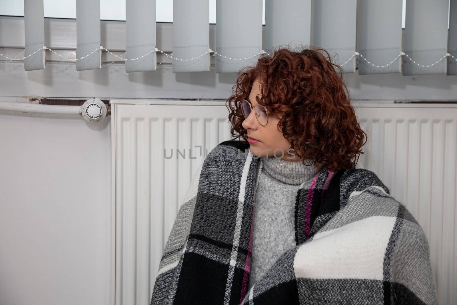 A woman sits on the floor with her back leaning against a radiator. The adult girl has a thick checkered scarf on her back. Central heating.