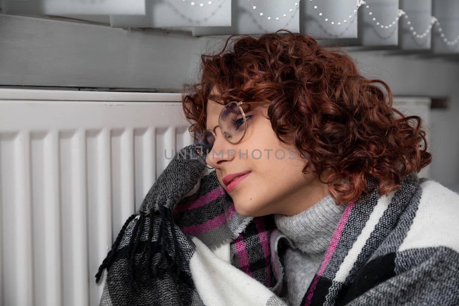Hot radiator. A girl with curly hair and glasses warms herself at the heater. Central heating during initial startup.