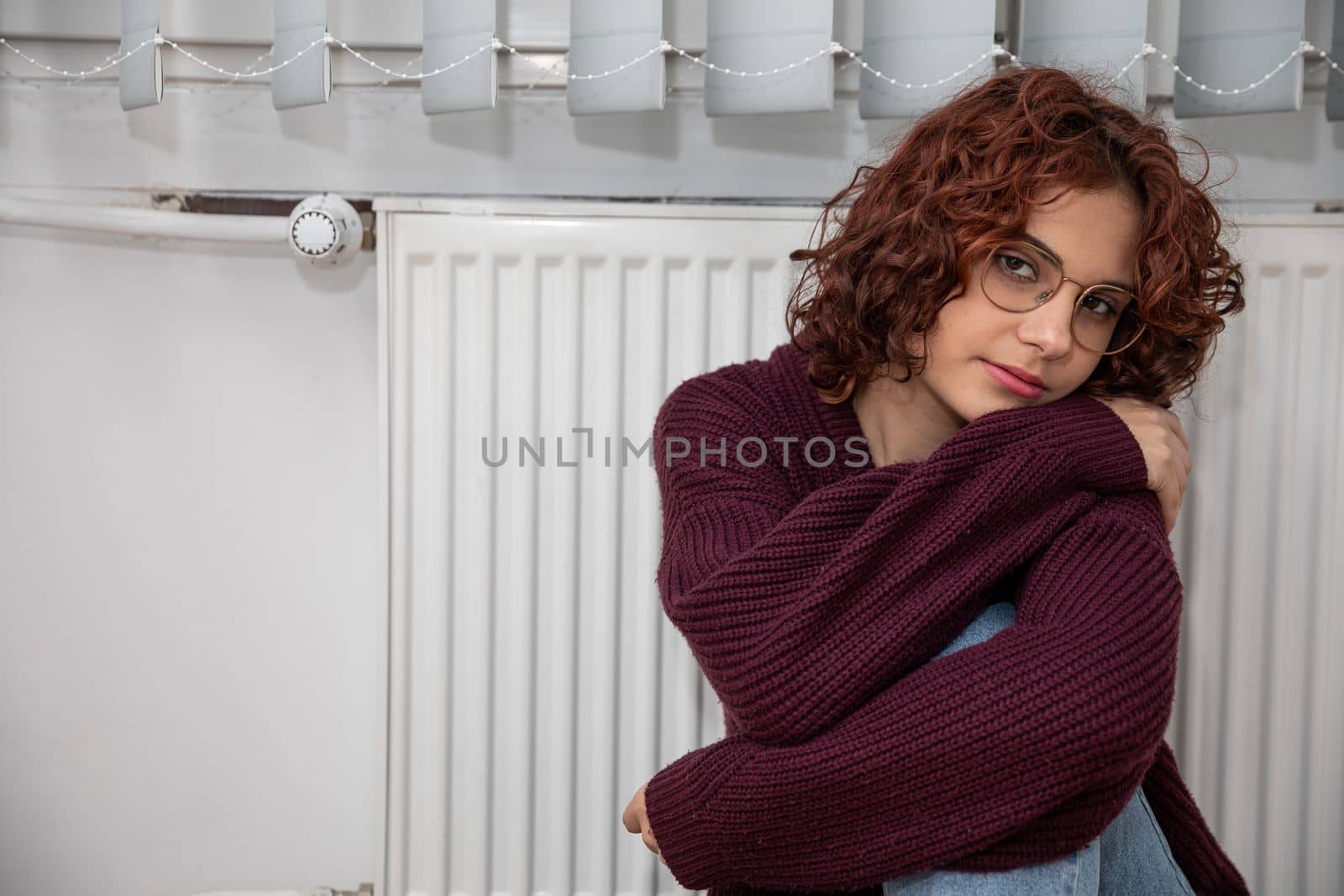 Dressed in a thick sweater, the girl sits on the floor by the radiator hoping that they will finally start the central heating.