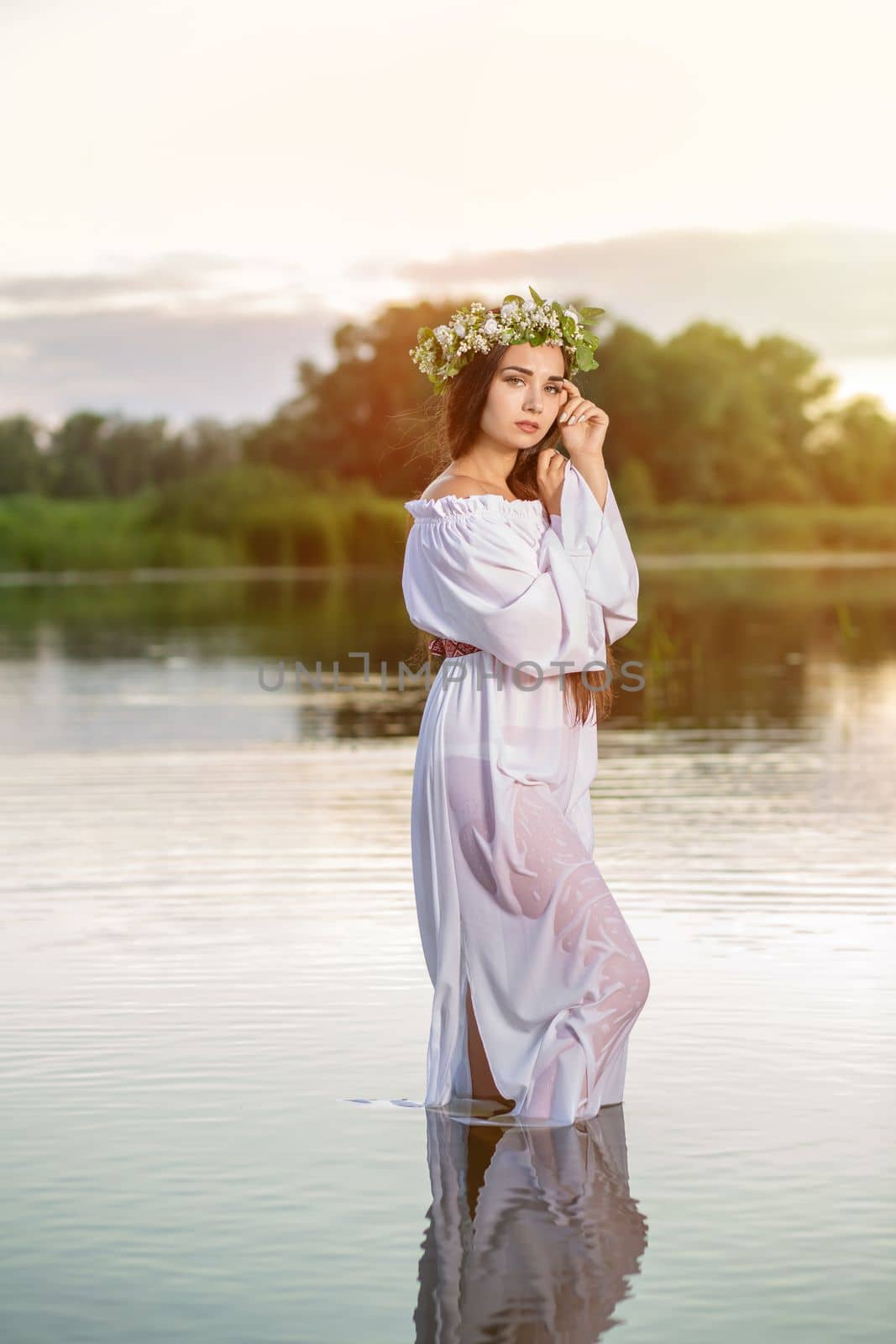 Beautiful black haired girl in white vintage dress and wreath of flowers standing in water of lake. Sun flare. by nazarovsergey