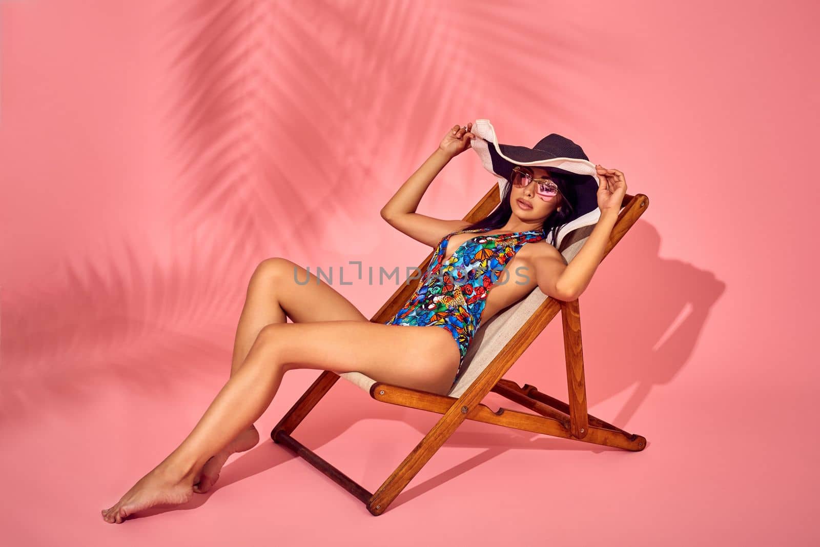 Summer lifestyle fashion portrait of young stunning woman on a lounge chair, pink background, studio shot. Enjoying life on the beach chaise. Wearing stylish sunglasses, bikini. Sunbathing.