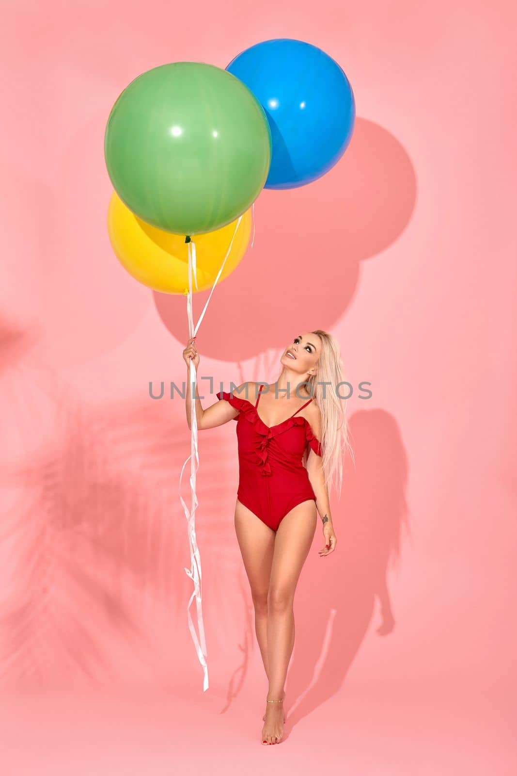 Young sexy slim woman in a red swimsuit is looking at balloons in her hand and posing in a studio. Full length fashion portrait of a beautiful girl with long blond hair, on a pink background.