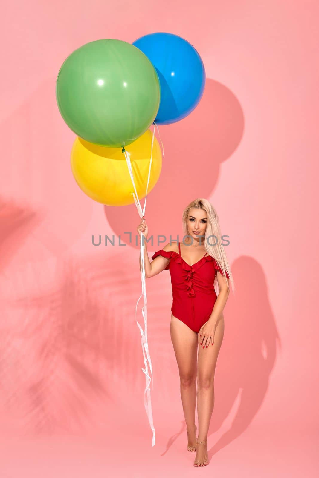 Young sexy slim woman in a red swimsuit with balloons in her hand is posing in a studio. Full length fashion portrait of a beautiful girl with long blond hair, on a pink background.