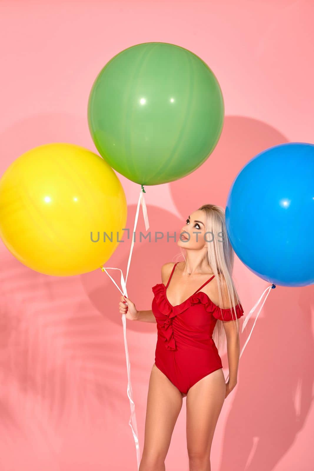 Young sexy slim woman in a red swimsuit with balloons in her hand is posing sideways in a studio. Full length fashion portrait of a beautiful girl with long blond hair, on a pink background.
