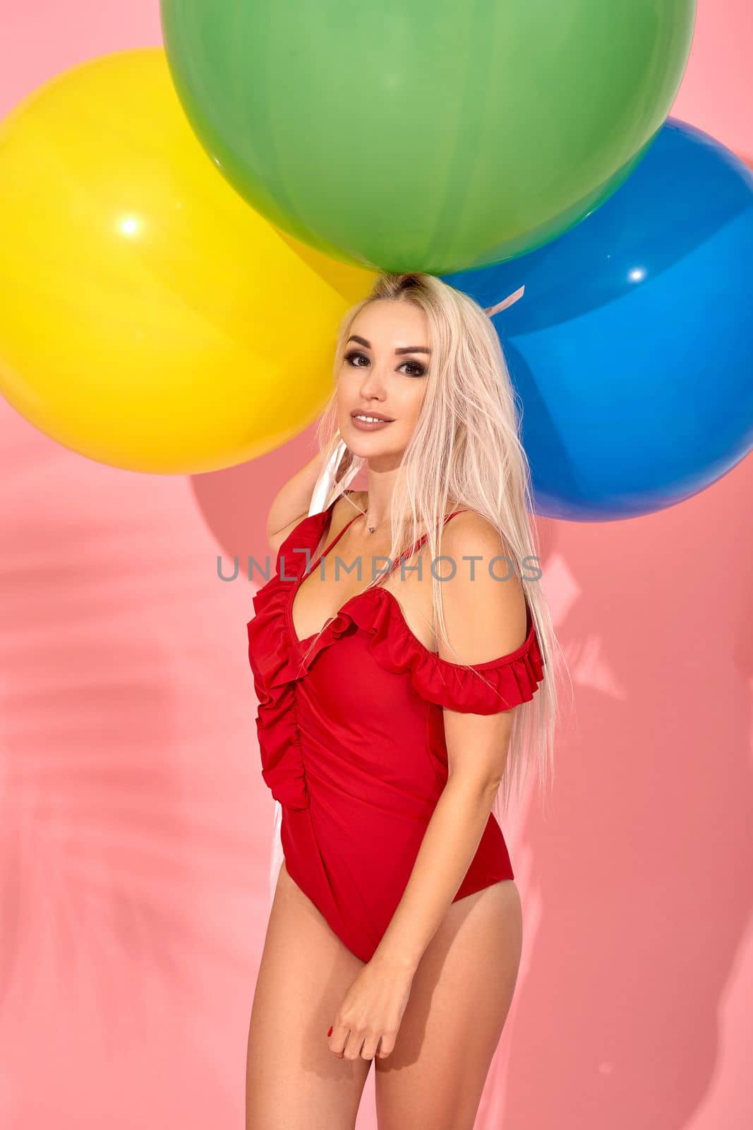Young sexy slim woman in a red swimsuit with balloons in her hand is looking at the camera. Full length fashion portrait of a beautiful girl with long blond hair, on a pink background.