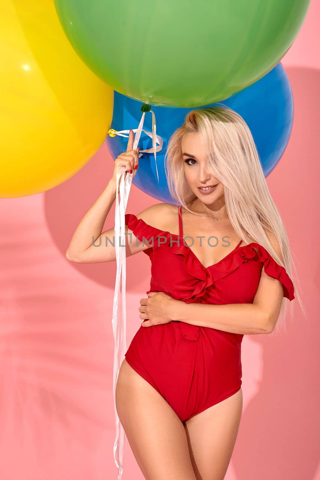 Young sexy slim woman in a red swimsuit with balloons in her hand is smiling at the camera. Full length fashion portrait of a beautiful girl with long blond hair, on a pink background.