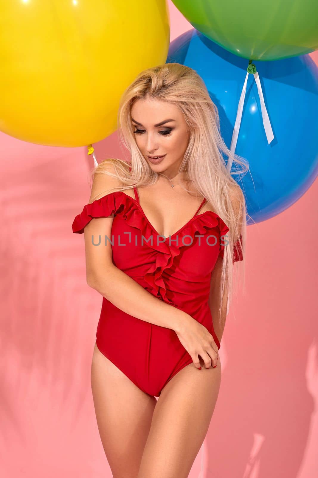 Sexy slim woman in a red swimsuit with balloons in her hand put her hand on the hip. Full length fashion portrait of a beautiful girl with long blond hair, on a pink background.
