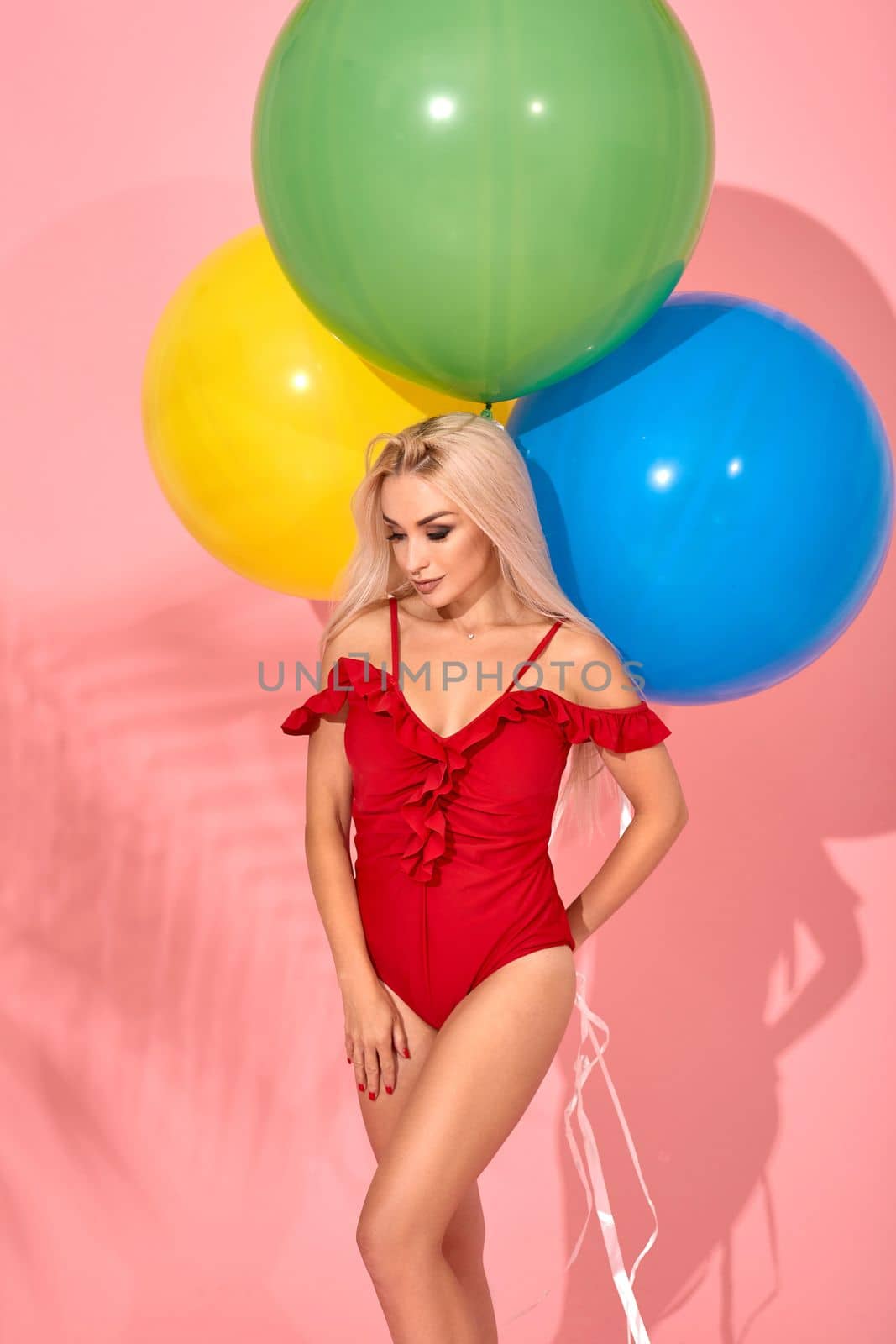 Sexy slim woman in a red swimsuit with balloons in her hand is looking down. Full length fashion portrait of a beautiful girl with long blond hair, on a pink background.