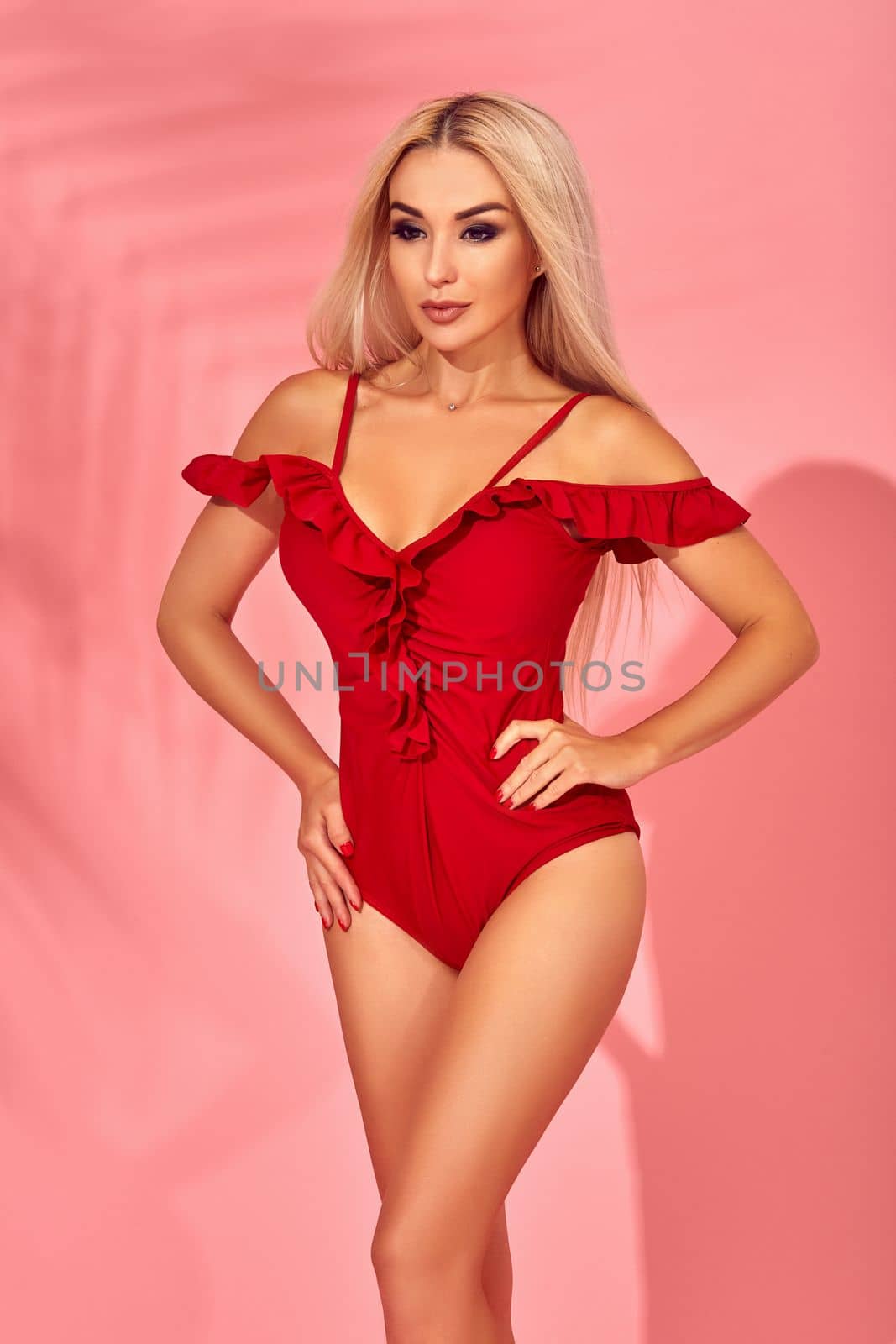 Stunning blonde female model with amazing body standing in an elegant red swimsuit on pink background with palm shadow by nazarovsergey