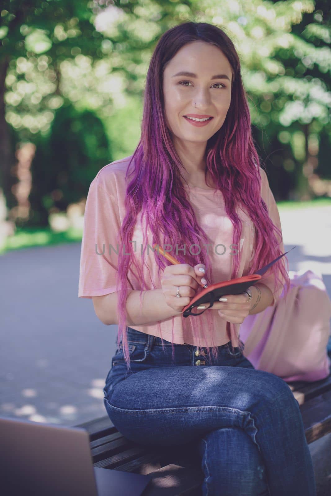 Woman writing in a notebook sitting on a wooden bench in the park. Girl working outdoors on portable computer, copy space. Technology, communication, freelance and remote working concept.