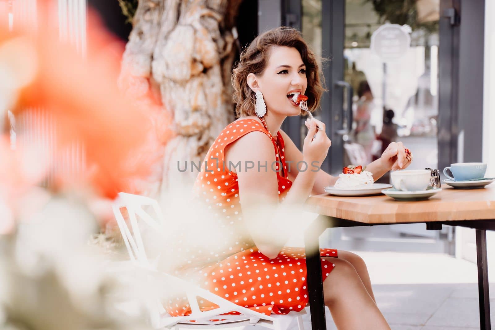 Charming woman in a restaurant, cafe on the street. She sits at the table and eats a cake with a fork. Dressed in a red sundress with white polka dots. by Matiunina
