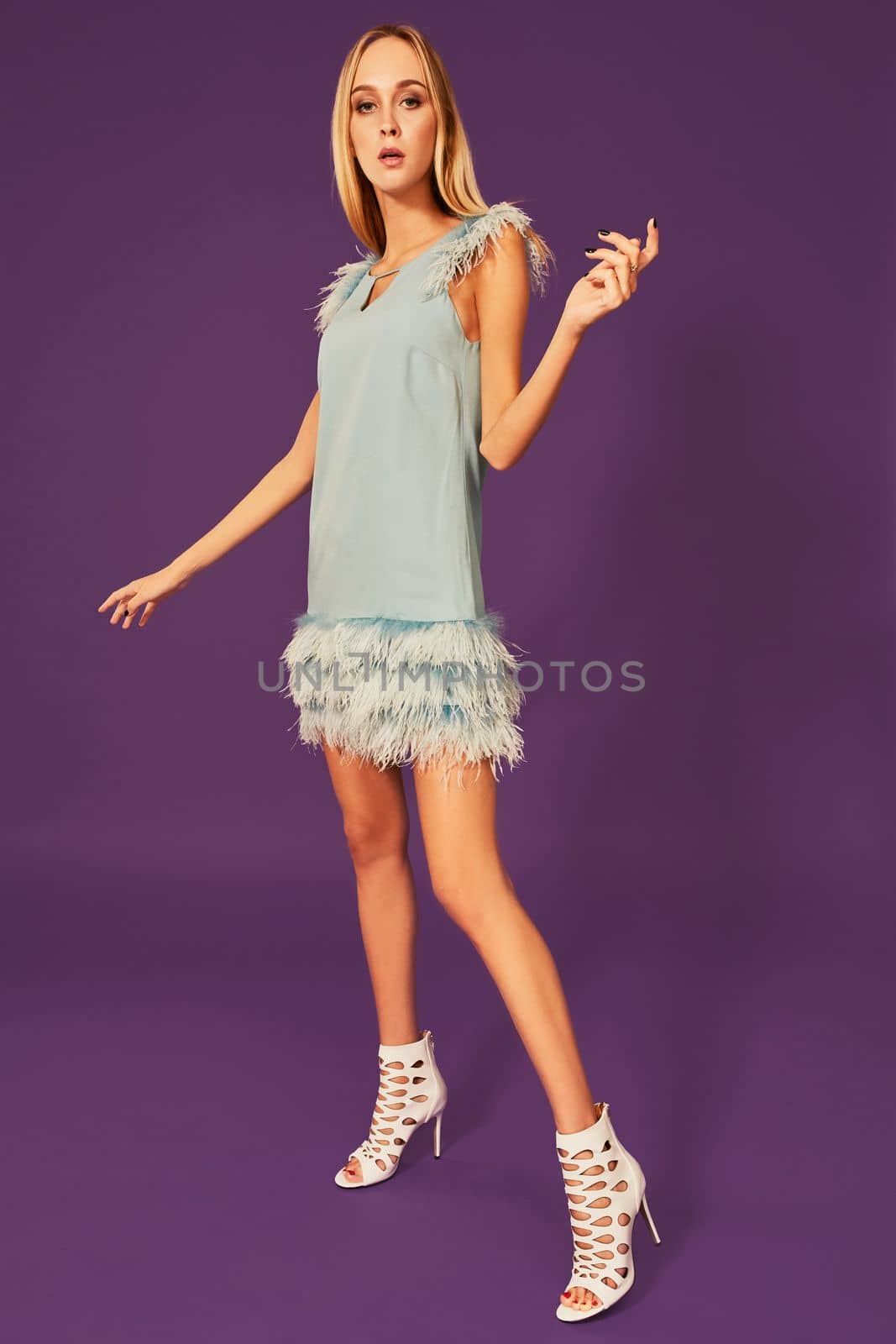 Young attractive woman with blonde long hair, dressed in elegant cocktail dress with feathers, wearing white sandals or shoes, posing at studio before camera