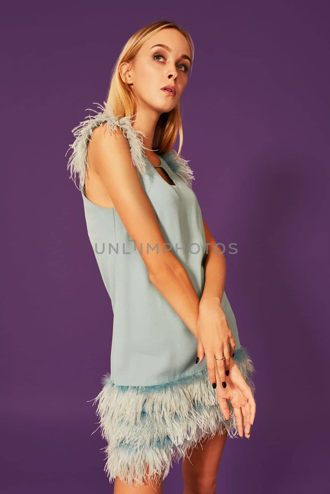 Young attractive woman with blonde long hair, dressed in elegant cocktail dress with feathers, posing at studio before camera with crossed hands