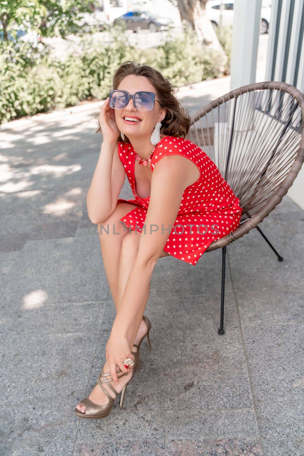 Portrait of a woman with glasses. Photo of a fashionable girl with beautiful brown hair, dressed in a red sundress with polka dots, sits in a chair on the street