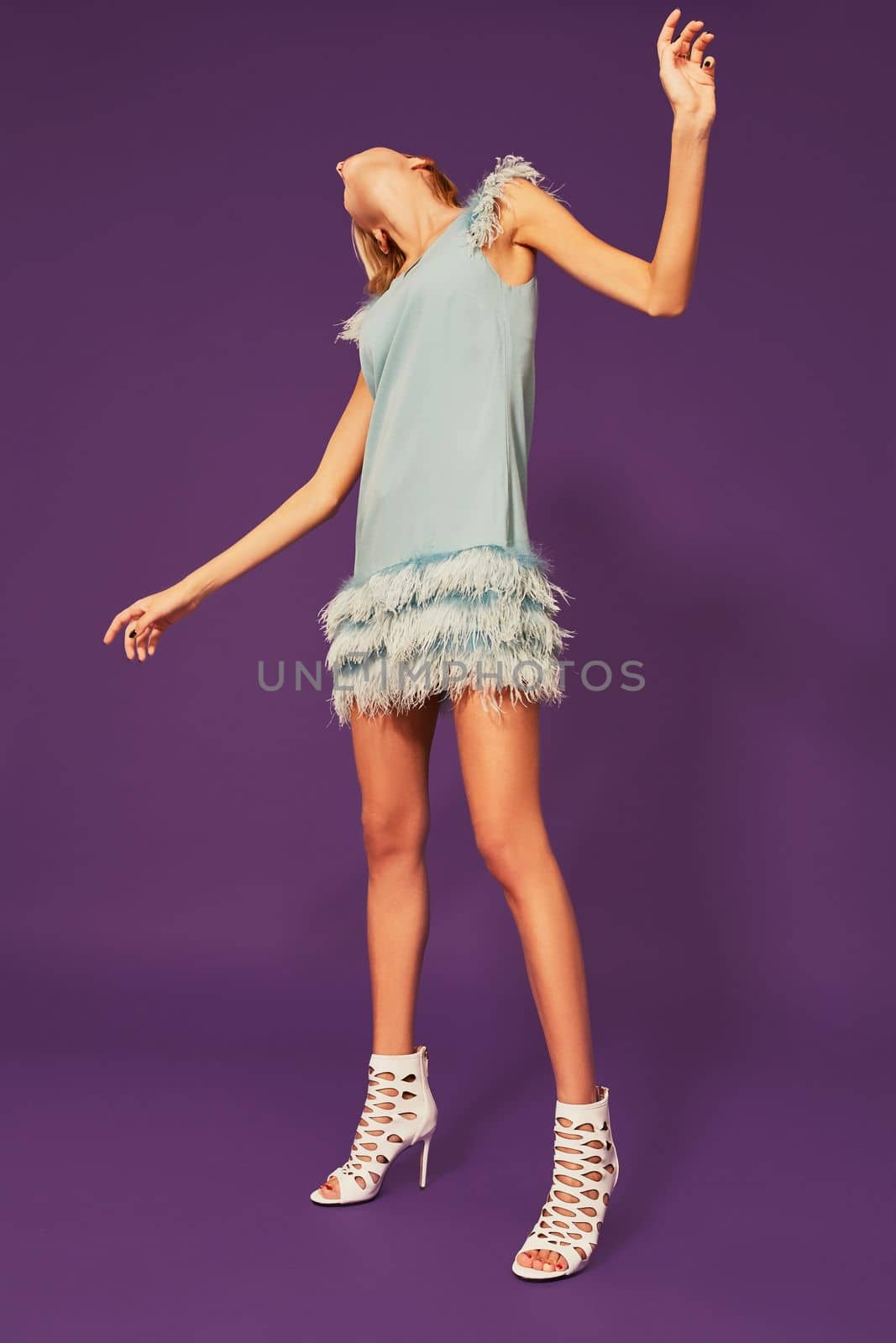 Photo of pretty young blonde girl in blue cocktail dress dancing on purple background in studio. Indoor portrait of caucasian young lady expressing her emotions