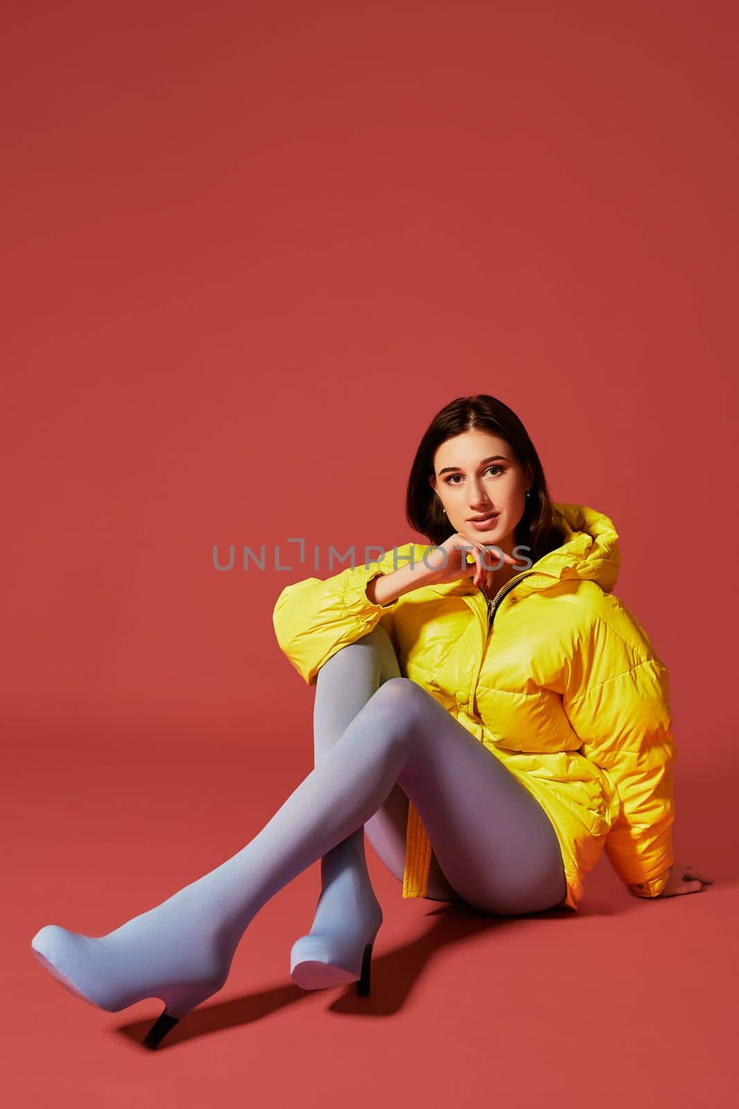 Young dark haired girl sitting on the floor before camera in studio on red background. Fashion image of stylish woman wearing yellow jacket