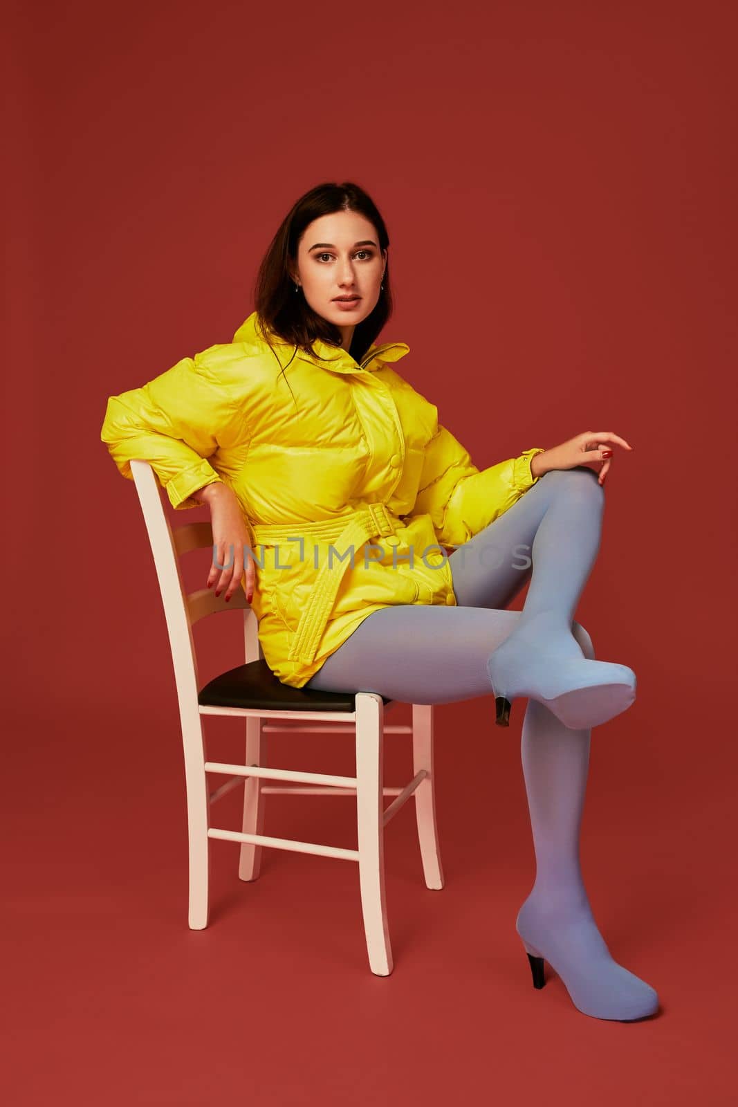 Stylish photo of cute brunette girl wearing yellow oversized down jacket posing in studio on red background, using chair