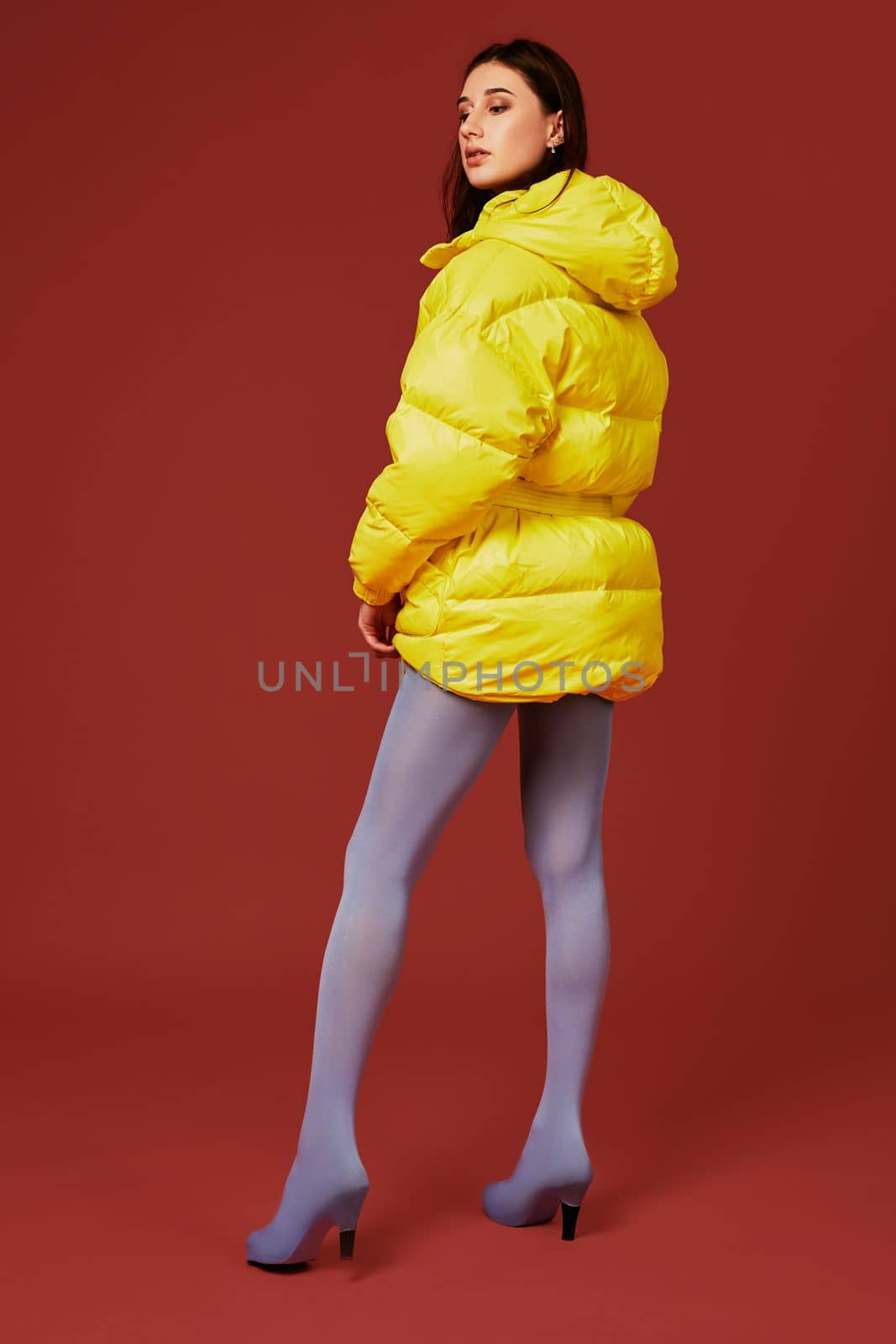 Image of young dark haired girl posing before camera in studio on red background. Fashion image of stylish woman wearing yellow jacket. Back view