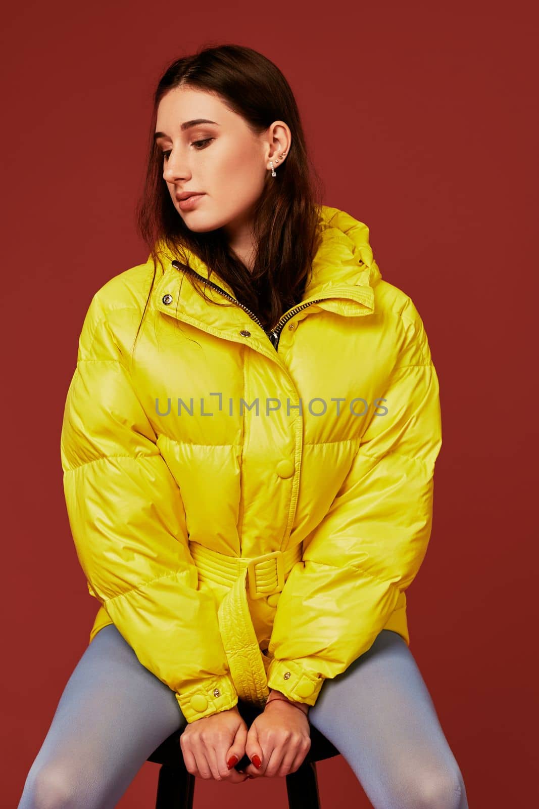 Close up stylish photo of cute brunette girl wearing yellow oversized down jacket posing in studio on red background, using chair