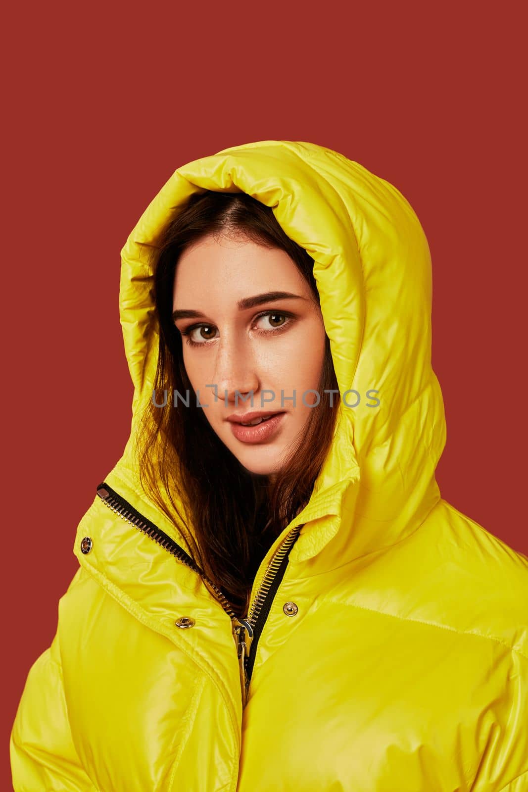 Close up photo of cute girl with dark hair in studio, in bright yellow down jacket. Red background, oversized clothes, hood on