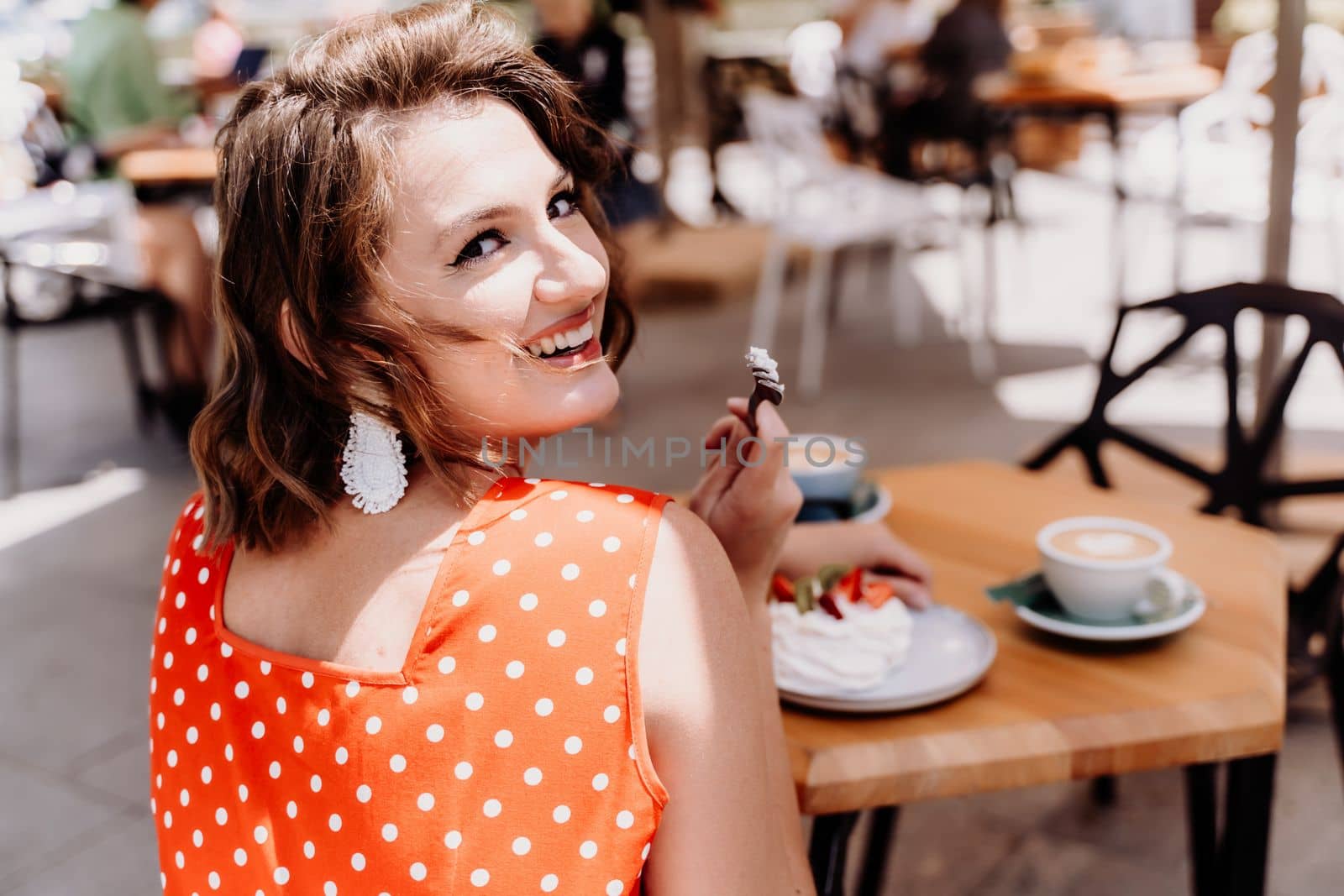 Charming woman in a restaurant, cafe on the street. She sits at the table and eats a cake with a fork. Dressed in a red sundress with white polka dots