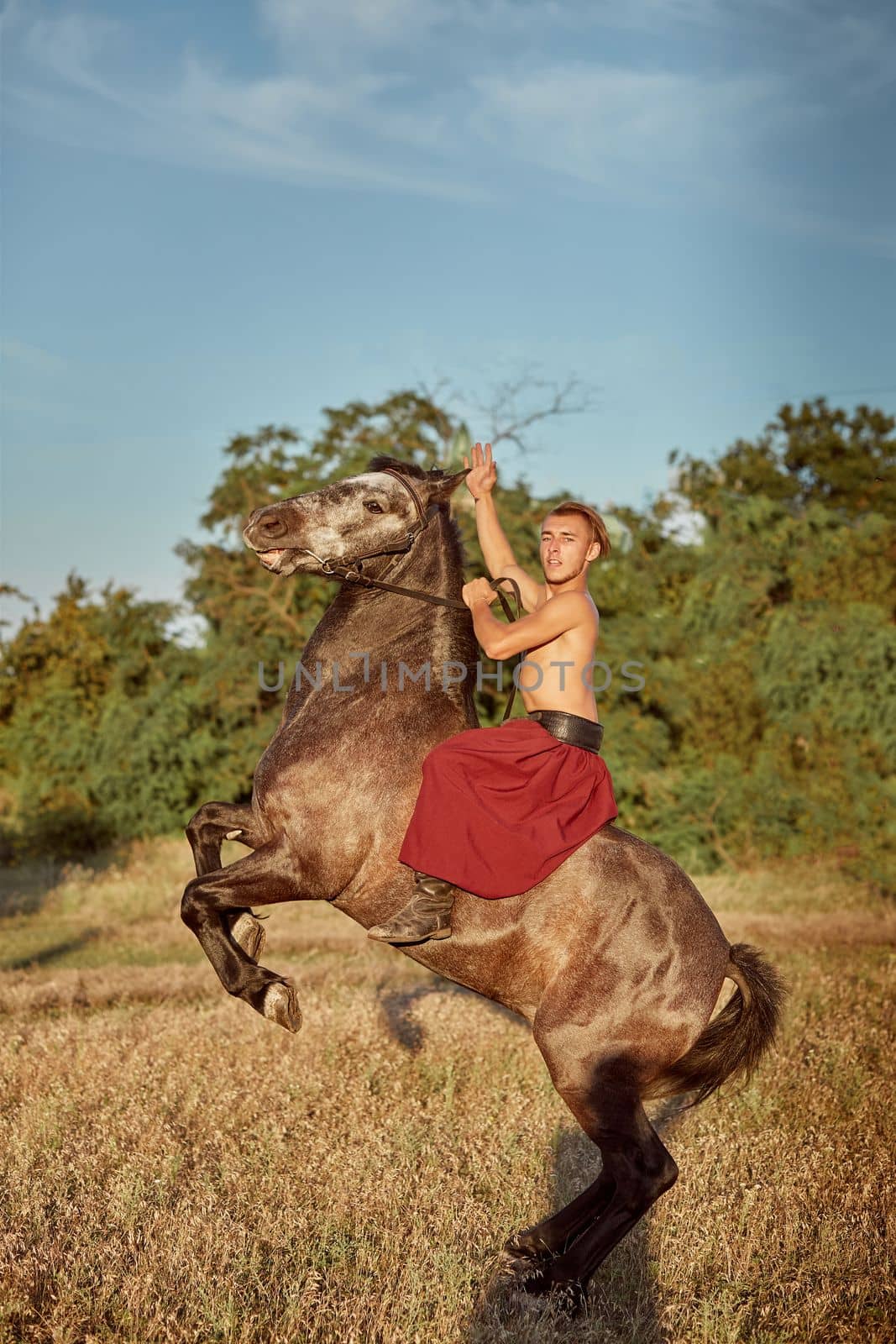 Handsome man cowboy riding on a horse - background of sky and trees. A man in red wide pants without a shirt. Show