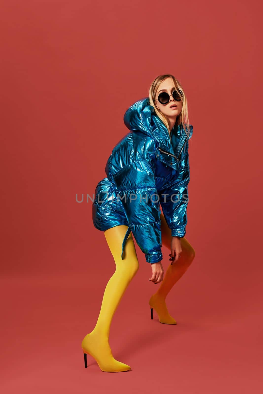 Fashion photo of emotional blonde girl posing on camera in bright blue warm jacket, sunglasses and yellow stockings. Oversized style