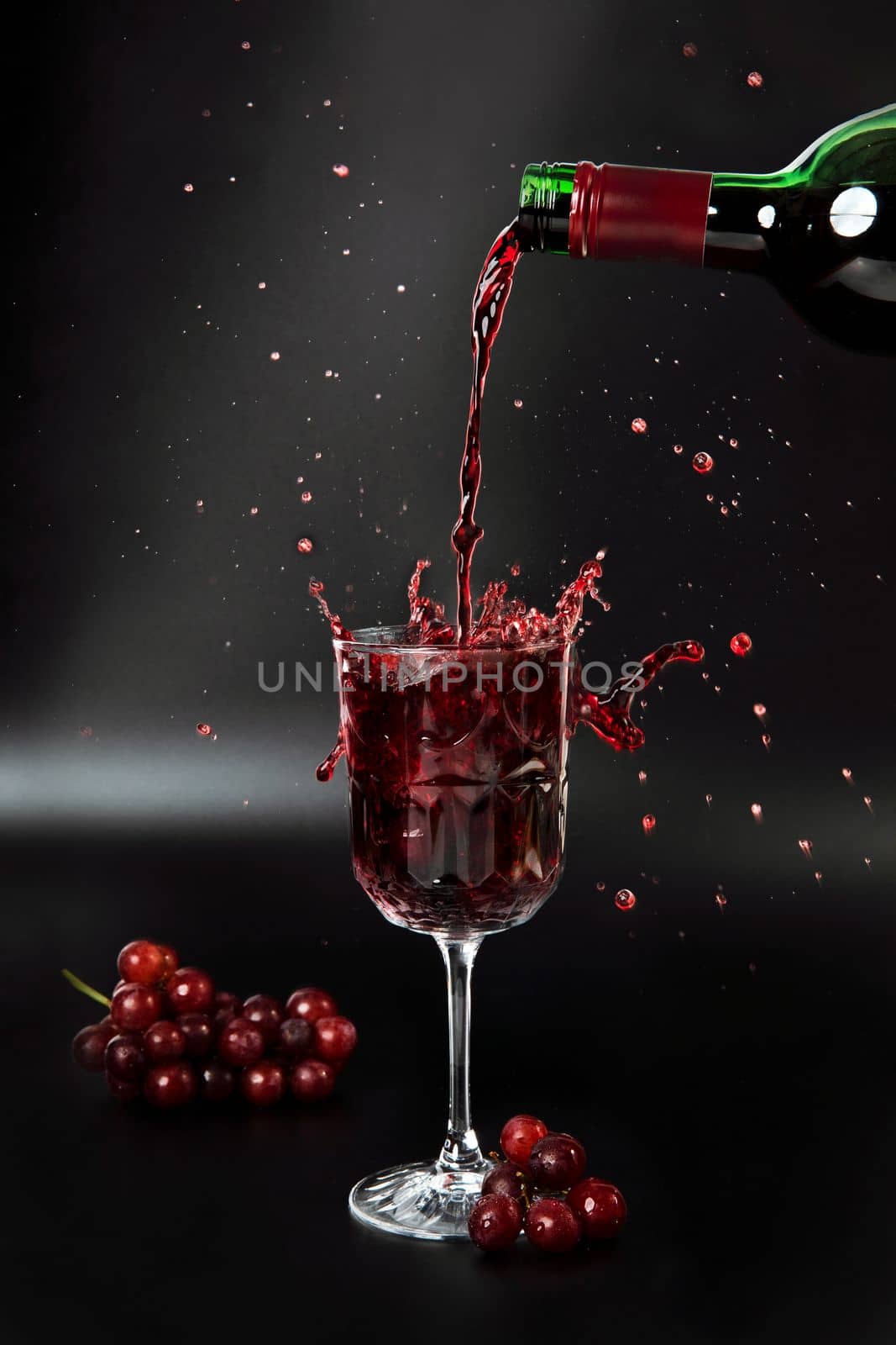 Red wine is poured into a wine glass on a black background with grapes and splash copy space