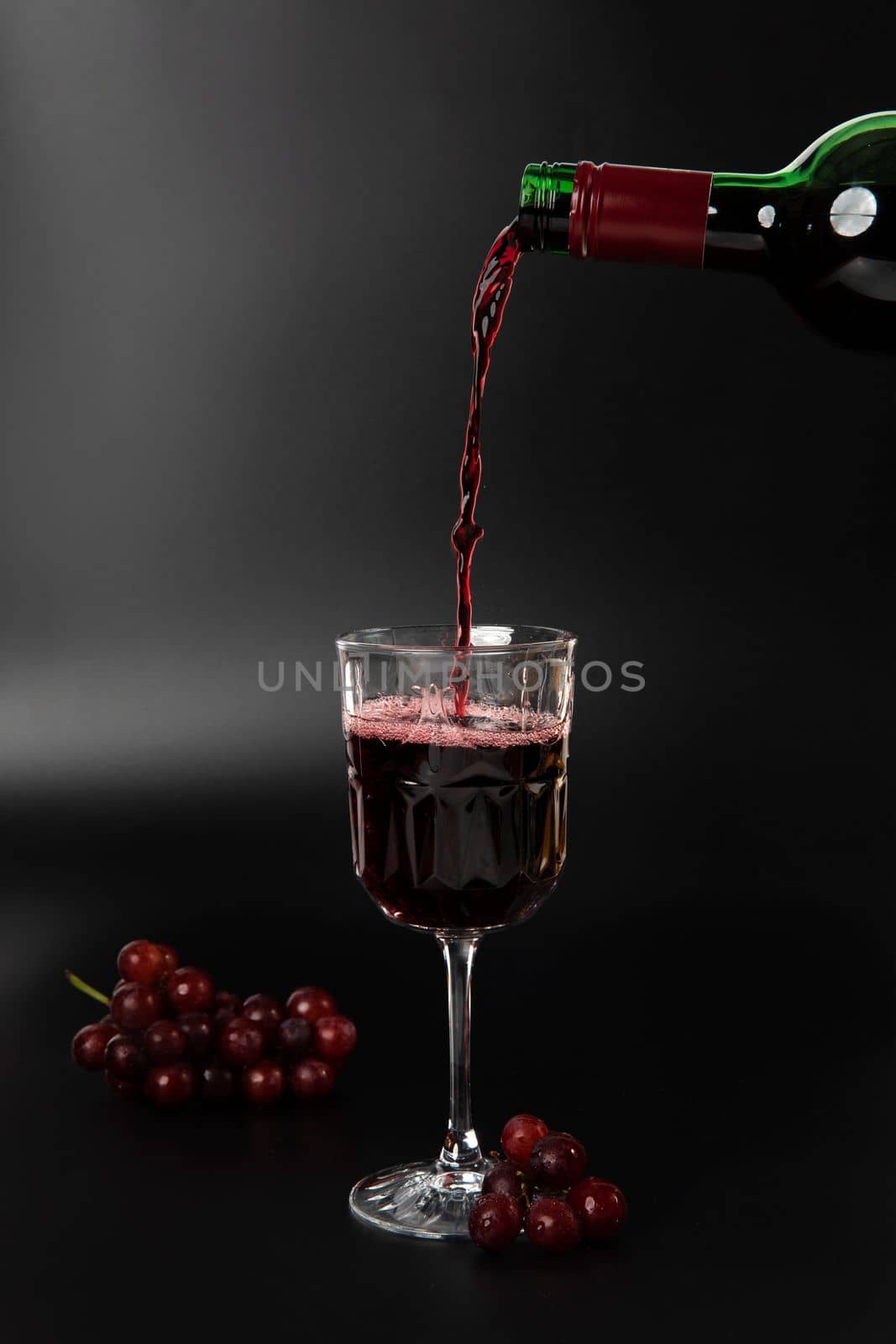 Red wine is poured into a wine glass on a black background with grapes and splash by Annebel146