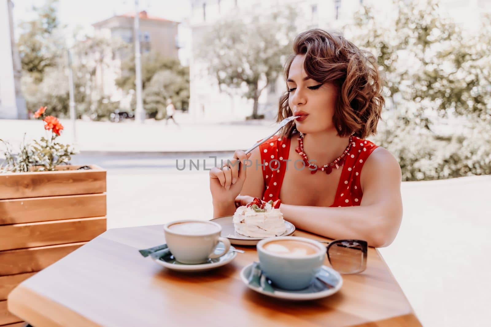 Charming woman in a restaurant, cafe on the street. She sits at the table and eats a cake with a fork. Dressed in a red sundress with white polka dots