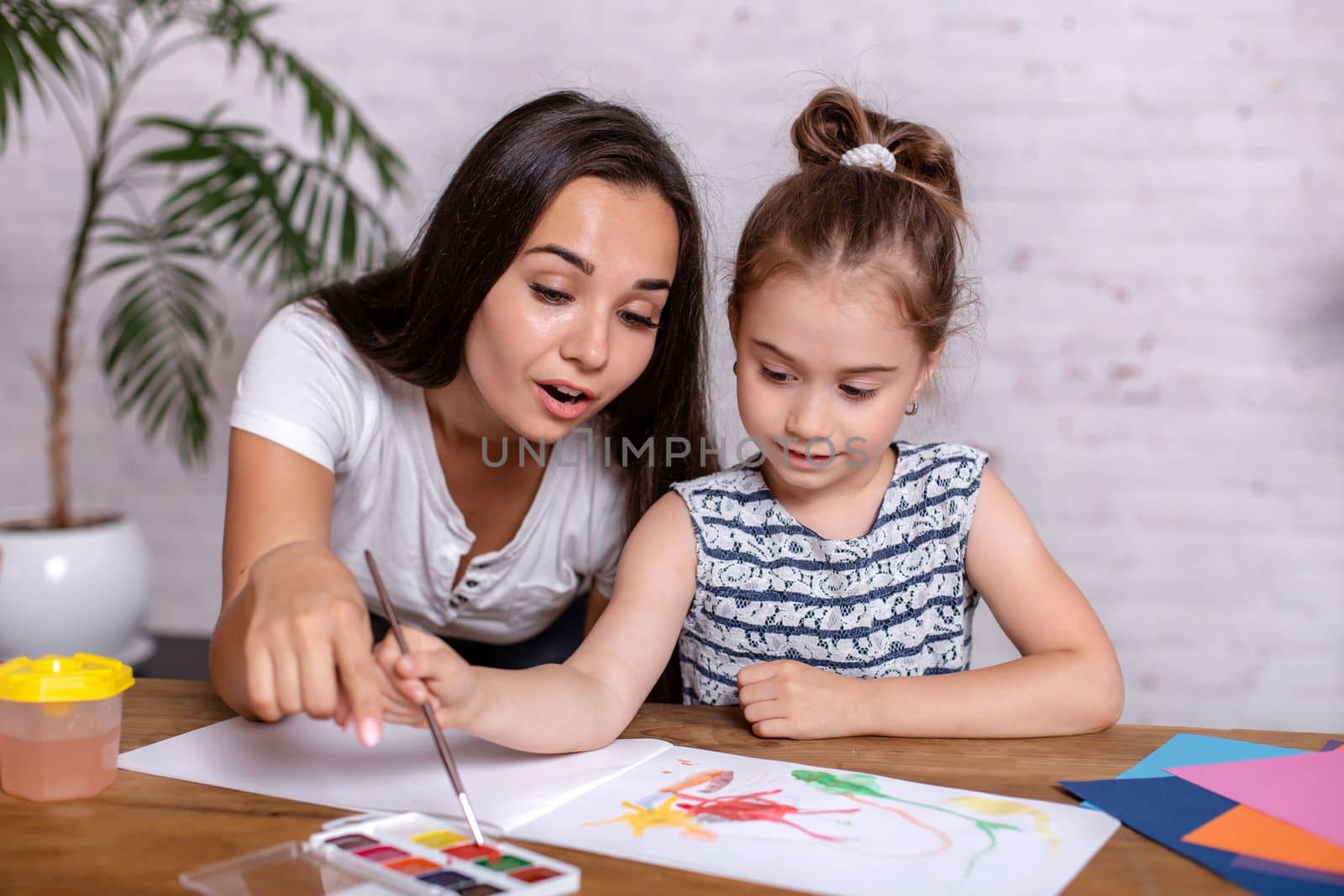 Attractive young woman and her little cute daughter are sitting at the table and having fun while doing homework together. Mother helps daughter with her school classes.