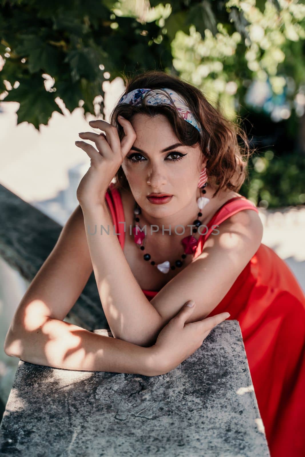 A pretty woman in a red silk dress and a bandage on her head smiles against the background of the leaves of a tree. She is leaning on the coop and looking into the camera. Vertical photo. by Matiunina